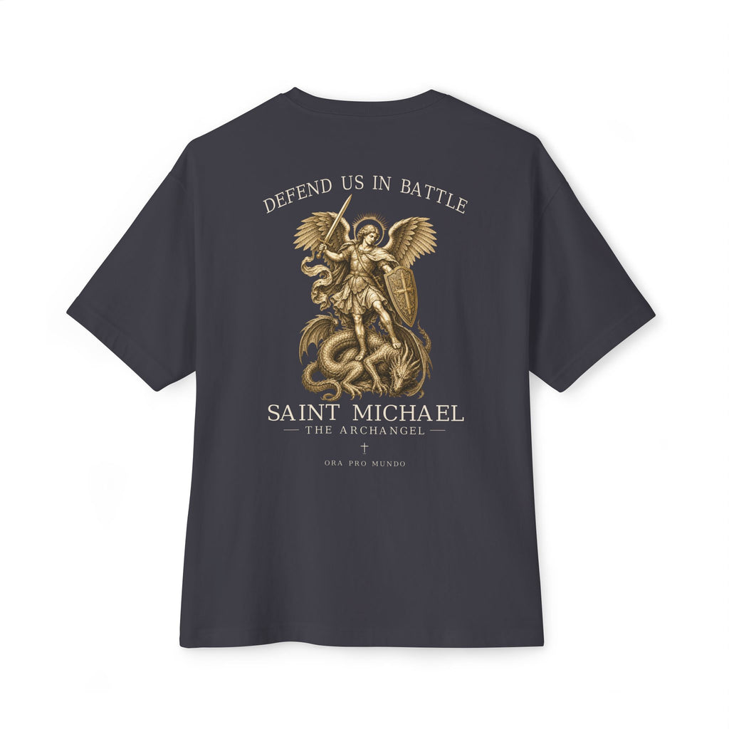 A dark charcoal gray t-shirt shown from the back, featuring a large gold graphic of Saint Michael the Archangel defeating a serpent. The text 'DEFEND US IN BATTLE' is printed above the graphic, and 'SAINT MICHAEL THE ARCHANGEL' is printed below.